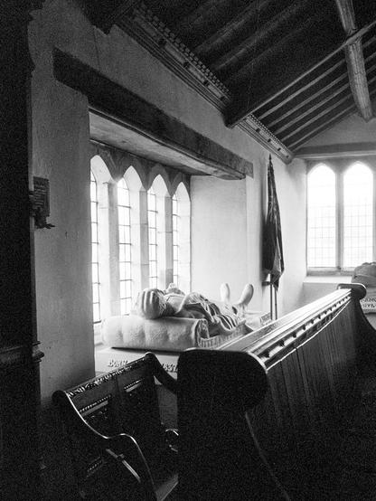 A rather dark portrait format black and white photo, lit by windows from the left and far end of a small chapel in a church building. A wooden pew and chair are jammed up against a stone tomb under the window, topped with an effigy of a figure, head nearest resting on a stone cushion. There is a flag between the two windows.