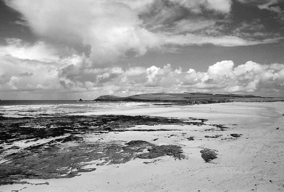 Black and white photo of a beach under broken fluffy white cloud. There are rocks to the left, and the beach stretches out on the right side towards a distant headland. There are few people on the beach, and the surf is rolling in, but at a distance. It is quite high contrast.