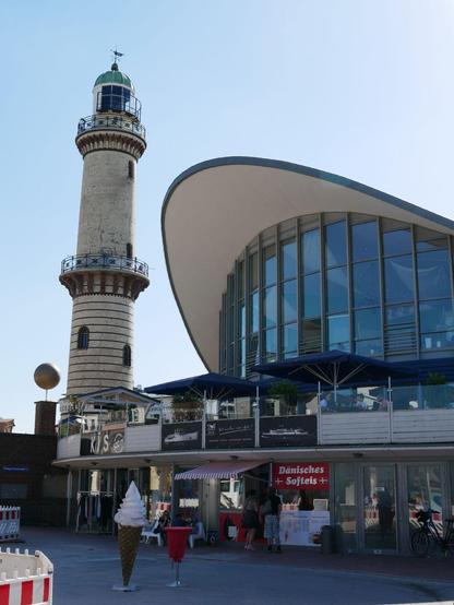 Blick auf den Warnemünder Teepott, einem Betonbau aus den 60ern mit rundem Dach und Glasfassade. Im unteren Geschoss sind kleine Geschäfte und ein Eisstand, das obere ist von einer Terrasse mit Tischen und Schirmen umgeben. Links angrenzend ist der Leuchtturm von Warnemünde zu sehen.