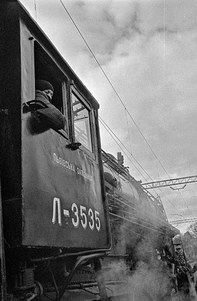 The image depicts a vintage steam locomotive, labeled with the number "Л-3535" on its side. The text above the number reads "Львівська залізниця" (Lviv Railway), indicating it belongs to the Lviv Railway network. A person, dressed warmly in a hat and coat, is seen leaning out of the cab window, looking outwards. Steam rises from the engine, adding to the industrial and historical atmosphere. Overhead, power lines and a cloudy sky are visible, giving the scene a nostalgic, black-and-white aesthetic.
