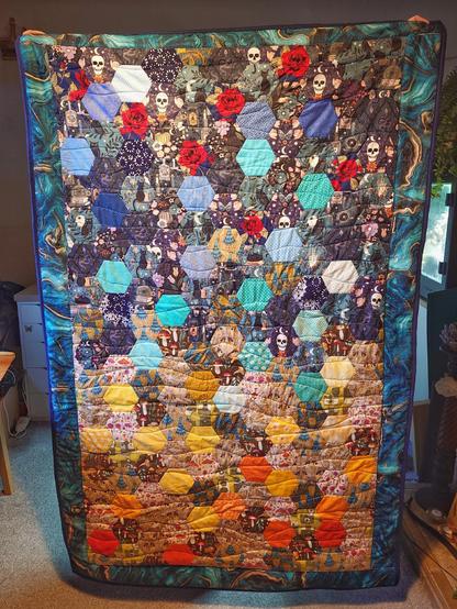 A large handmade quilt being held up in front of a person. It's made of hexagons, blue at the top and yellow at the bottom. It's bordered by a marbled blue and gold fabric, with a navy blue binding around the edges. 