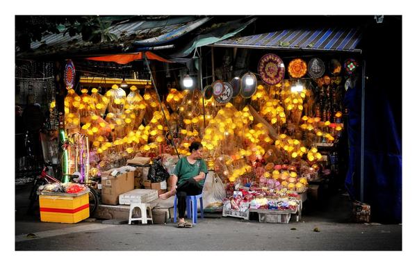 This image depicts a vibrant street-side shop at night, illuminated by an array of glowing yellow lights that resemble flowers or lanterns. The shop is densely packed with these light fixtures, giving it a warm, golden glow. A woman, dressed casually in a green top and black pants, is seated on a small blue plastic stool outside the shop. She appears relaxed and is looking away, perhaps observing her surroundings. The shop also displays various other items, including boxes, baskets, and decorations, adding to the bustling atmosphere of the scene. The setting seems to be a local market or street vendor area, with other stalls partially visible in the background.
