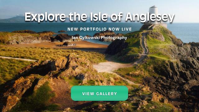 The image shows a stunning coastal landscape of the Isle of Anglesey, with rugged cliffs, grassy hills, and a picturesque lighthouse perched on a rocky outcrop overlooking the sea. The text overlay reads, "Explore the Isle of Anglesey," followed by "New Portfolio Now Live" and "Ian Cylkowski Photography." A green button at the bottom says "View Gallery." The scene is bathed in warm sunlight, highlighting the natural beauty of the location.