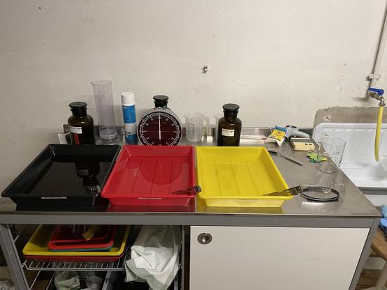 A workstation with three plastic trays (black, red, and yellow) containing liquids and arranged on a metal table. Visible items include glassware, measuring equipment, chemical bottles, a timer, and safety glasses. The background shows a sink and various supplies