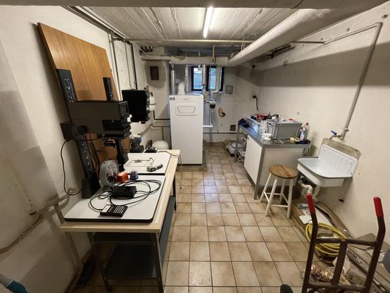 An interior basement room featuring a work table with a two enlargers, a small analogue timer, and various equipment. On the right, there is a washing machine and a utility sink. The floor is tiled, and the walls are painted white.
