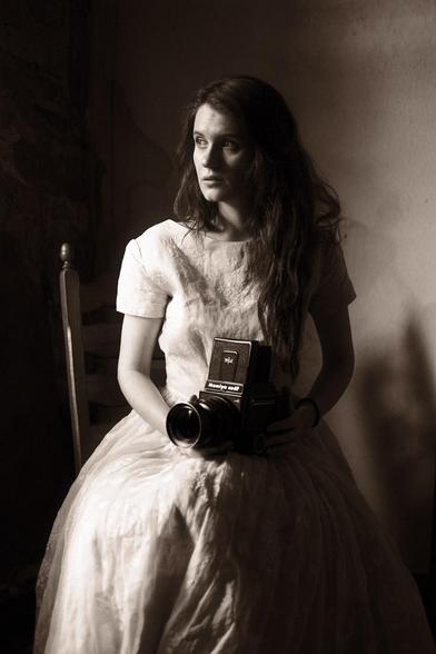 Young woman in an old white dress holding a Mamiya RB67 in her hand and looks out an off-screen window