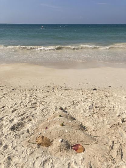 On a white sandy beach a Christmas tree is made out of sand and decorated with some coconuts and leaves. The blue ocean is in the background.