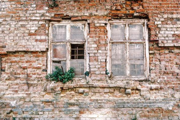 This image depicts a weathered brick wall with two old, worn-out windows. The window frames are made of wood and appear to be deteriorating, with peeling paint. One of the windows has a partially broken pane, and there is some greenery, possibly small plants or ivy, growing at the base of the wall and between the bricks. The structure gives off an abandoned or aged aesthetic, with the bricks showing various shades of red and gray due to wear and time.