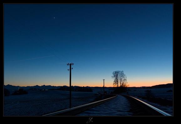 The image in landscape format was taken standing on a railroad track. The rails run left and right in a gentle left-hand curve into the distance. To the left of the track, two old style telegraph poles and wires can be seen silhouetted against the sky. A tree in the distance to the right of the tracks, the mountains on the horizon and a forest are just silhouettes. The sky changes from a strong orange above the horizon to a dark blue. Venus can be seen high up in the dark blue sky.


AI disclaimer: Using my work, its meta data, written or derived description to create media with or train AI based systems is prohibited.