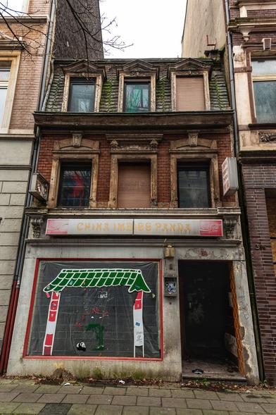Kleines,schmales und altes Haus aus Ziegelsteinen mit drei Fenstern im ersten Stock sowie drei Fenster im Mansardendach. Alle Fenster haben Sandsteingewände. An zwei Fenstern sind die Rollläden hinunter gelassen. Im Erdgeschoss ein verlassener Chinaimbiss mit einem Schaufenster. Direkt daneben der Eingang und darüber ein verblichenes Werbeschild fast über die gesamte Hausbreite.