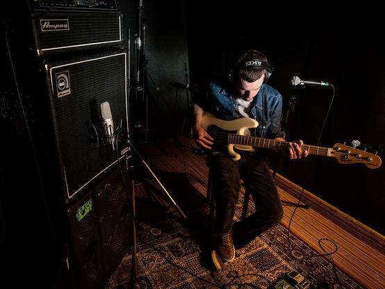 Aufnahmestudio. Ein Bassist sitzt rechts im Bild und spielt gerade auf seinem Bass, links der Verstärkerturm, vor dem ein Tonabnehmermikrofon steht.