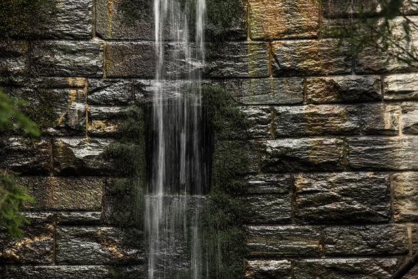 Ein kleiner Wasserfall läuft eine gemauerte Wand hinunter. Hinter dem Wasser erkennt man eine rechteckige dunkle Öffnung in der Mauer.