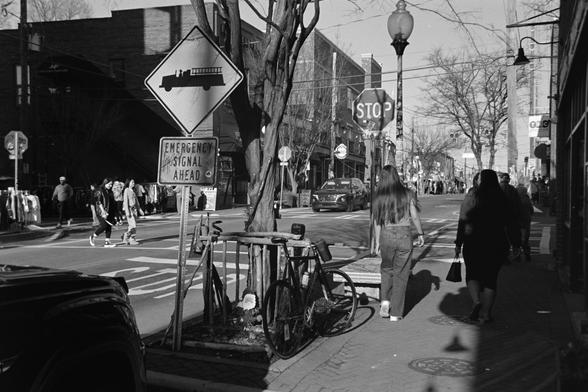 A view of a street corner; people walking in multiple directions, a rustic bike rack that looks like it’s made of branches of wood