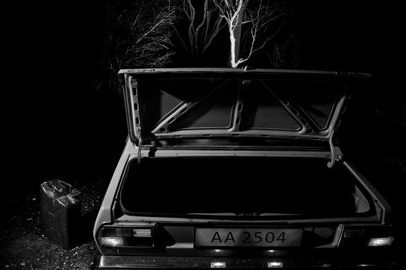 Wir sehen den leeren Kofferraum eines alten Audi 80 mit dem Kennzeichen AA2504 und dem Länderkennzeichen (noch als Aufkleber und nicht mit dem Nummernschild) "L" für Luxemburg. Neben dem leeren Kofferraum steht ein Benzinkanister.

Das "L" weist also eben auf die Herkunft aus Luxemburg hin. Zur Zeit dieses Autos (ein Modell der 70er Jahre) hatten Behördenfahrzeuge in Luxemburg Kennzeichen, die mit AA anfingen. Leicht zu übersehen und kaum einer weiß es halt ... :-/

Ursprünglich lag hier im Kofferraum noch eine gefesselte Person, aber der Dozent meinte, dass man sie nicht in jedem Bild sehen müsse und dass das folgende Bild reichen würde, um diesen Part der Geschichte zu erzählen, deswegen ist sie hier rausgeshoppt worden.