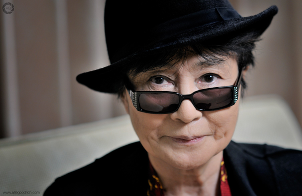 Yoko Ono wearing a black felt trilby style hat. She's also wearing slim, rectangular shaped sunglasses, which are studded with rhinestones on each side. She's smiling, looking directly at camera. The back of a small, low sofa is in the background out of focus. 