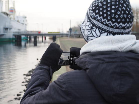 Einem Menschen beim fotografieren mit dem Handy über die linke Schulter fotografiert. Der Mensch trägt eine warme dunkle Jacke, schwarze Handschuhe und eine Schwarzweiße gemusterte Mütze. Der Fokus vom Bild ist das Handy, auf dem die Kamera-App mit dem Motiv, einem großen Schiff und dem Uferbereich zu sehen ist.
Im Hintergrund des Bildes sind ein Teil des Schiffs sowie das Ufer und die Stege unscharf zu sehen.