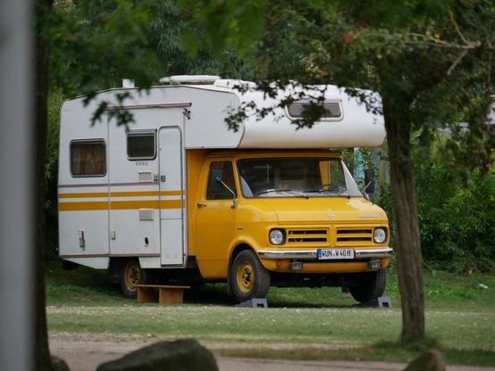 Ein Wohnmobil basierend auf einem alten Opel Transporter (zwischen 1996 und 1980 gebaut) steht zwischen Bäumen. Das Auto ist Senfgelb lackiert, der Aufbau ist weiß mit zwei horizontalen Streifen in Wagenfarbe. Vor der Tür steht ein Holzbänkchen als Trittstufe.