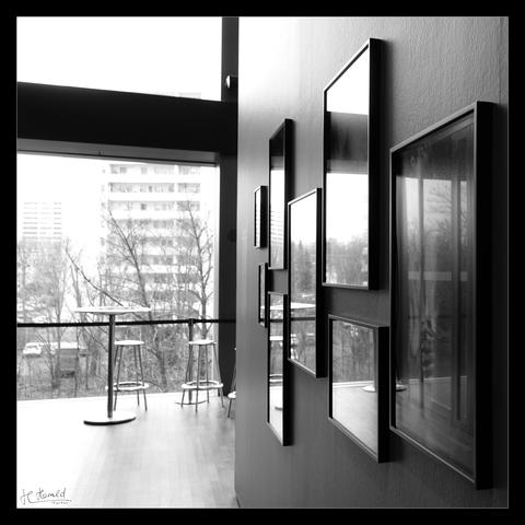 Square black and white picture of a photo exhibition with black frame. In the right half of the picture is a grey wall photographed at a shallow angle with 8 photos of different sizes behind glass in thin frames. The photos are not recognisable because the daylight is reflected in them. The light falls in through large windows on the left of the picture. The window front is divided by a massive horizontal bar made of metal. There are metal stools and a bar table in front of the window and a barrier tape is stretched in front of it. Outside you can see leafless trees, tower blocks and cars.