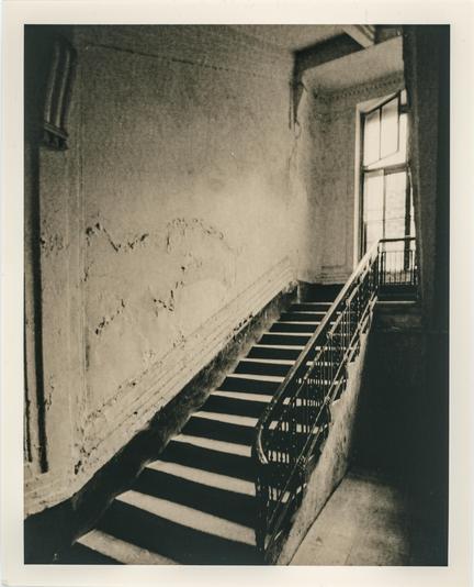 The image depicts an old, abandoned staircase in a dimly lit building. The stairway has a metal railing with intricate details, and the walls show signs of decay, with peeling paint and cracked plaster. A large window at the top of the stairs allows some light to enter, casting dramatic shadows on the steps and emphasizing the textures of the worn surfaces. The composition has a strong contrast, characteristic of the lith print technique, which enhances the gritty, timeless, and atmospheric feel of the scene. The grainy, high-contrast effect gives the photograph a vintage, moody aesthetic, emphasizing the passage of time and abandonment.