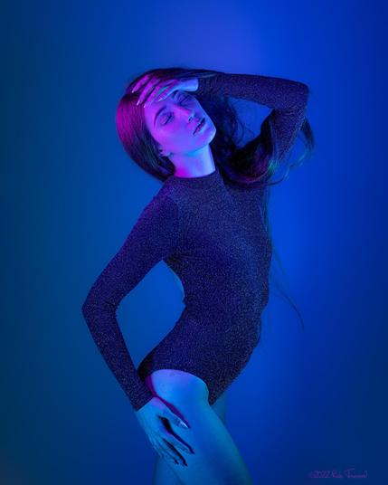 A photo of art model Daria S by photographer Rab Townsend. Daria is posing in a sparkly black bodysuit in blue and purple light. Only her face is cast in purple light. Her body is facing the right of frame as her right hand rests on her right thigh, and her left arm is raised so that her left hand is just in front of her face at her hairline. Her eyes are closed, and her red hair is tangled around her arm.