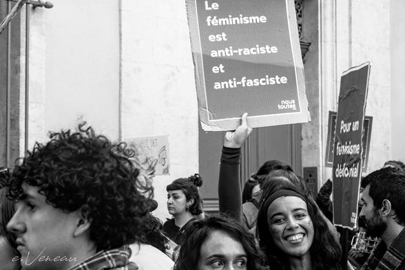 A smiling girl looks at the camera and holds up the sign "Feminism is anti-racist and anti-fascist".