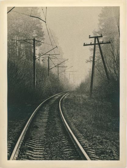 The image is a black-and-white lith print created using an optical printing technique in a darkroom. It is printed on old photographic paper, giving it a vintage aesthetic. The photograph depicts a railway track curving gently into the distance, surrounded by dense trees and vegetation. Overhead electric lines and utility poles line the track, disappearing into the misty background, creating a sense of depth and mystery. The grainy texture and high contrast of the lith printing technique enhance the dramatic and atmospheric quality of the scene. The faded borders and slightly aged appearance of the paper contribute to the nostalgic feel of the print.