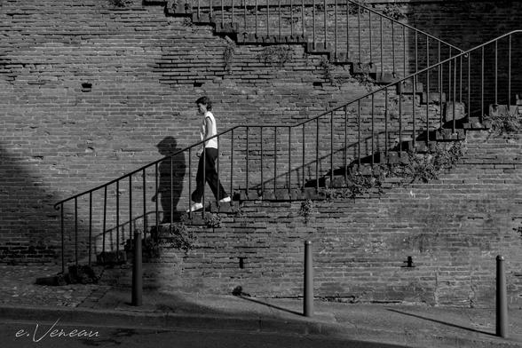 On the street, a sunlit brick wall supports two sets of stairs. A person is coming down it, in profile, wearing black pants and a white T-shirt. His shadow is on the wall.