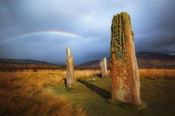 A serene and ancient stone circle at Machrie Moor on the Isle of Arran, Scotland. Three tall standing stones dominate the foreground, their surfaces weathered and covered with patches of lichen and moss, indicating their age and exposure to the elements. The largest stone stands prominently on the right, its rugged texture and earthy tones contrasting with the surrounding landscape. The other two stones are slightly smaller and positioned further back, creating a sense of depth and alignment. The ground is a mix of grass and dry, golden-brown vegetation, suggesting an autumnal setting. In the background, rolling hills stretch across the horizon under a partly cloudy sky, with a faint rainbow arching gracefully above the scene. The overall atmosphere is one of tranquility and historical significance, evoking a sense of connection to the past amidst the natural beauty of the Scottish countryside.