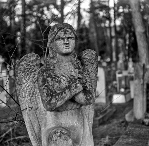 The image is a black and white photograph of an aged stone angel statue in a cemetery. The angel has a grim expression, with arms crossed over its chest and a small cross held in its hands. The wings are intricately carved but appear worn due to time and weathering. The statue is covered in patches of moss and lichen, adding to its aged and eerie appearance. In the background, blurred tombstones and tall trees create a mysterious and somber atmosphere, enhancing the sense of solitude and timelessness in the scene.