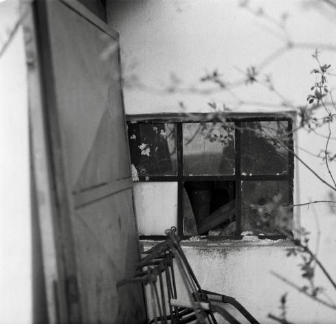 Black and white film photo. An old window with iron frame with six panes. Four panes with glass, one with the white plywood and one is empty. Some old iron door is leaning against the wall on the left side