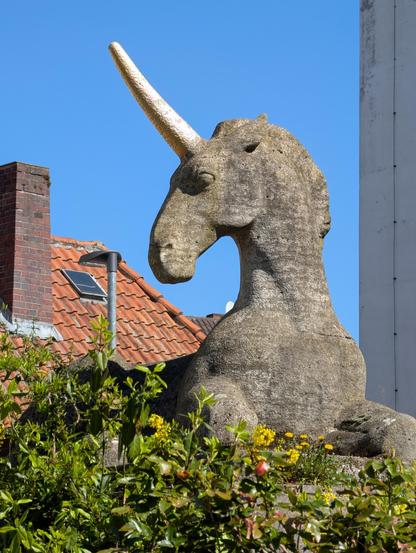 Eine Steinskulptur von einem liegenden Einhorn mit aufgerichtetem Kopf. Im Hintergrund sind vor dem blauen Himmel ein Hausdach und die Ecke eines Hauses zu sehen, davor einige grüne Büsche.