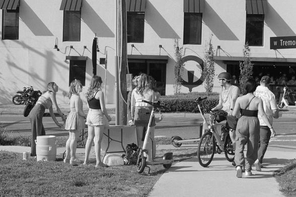 Young people grouped together at a street corner, some appear to be selling something at a table