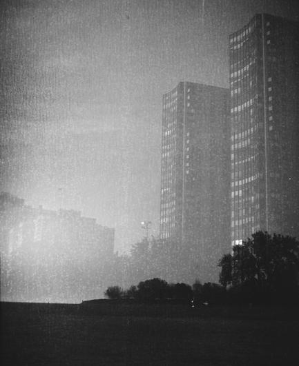 A grainy, slightly damaged looking black and white photo of two high-rise apartment buildings in Chicago on a foggy day. A beach with some trees is silhouetted against the buildings at the bottom of the image.