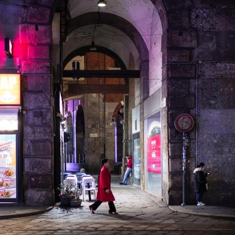 A slightly busy street scene with a man laying against a wall under a large arch, somewhat in the backgrdound, another man with a bright red coat walking past the arch, and a woman standing a one edge of the arch using her phone.