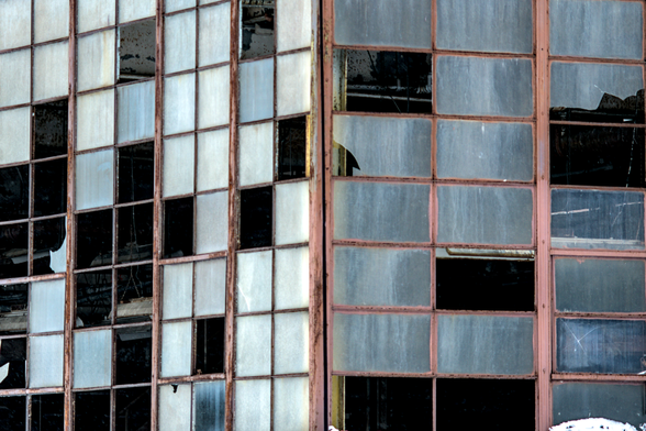 Factory window detail with rust and damage – Rows of dirty, broken glass windows with rusted metal frames show machinery and pipes inside the deteriorating building.