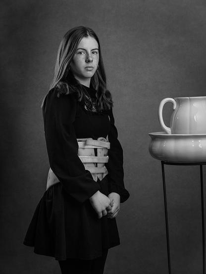 Studio portrait of a 12 year old girl in a black dress with a scoliosis back brace is standing next to a porcelain wash bowl and jug