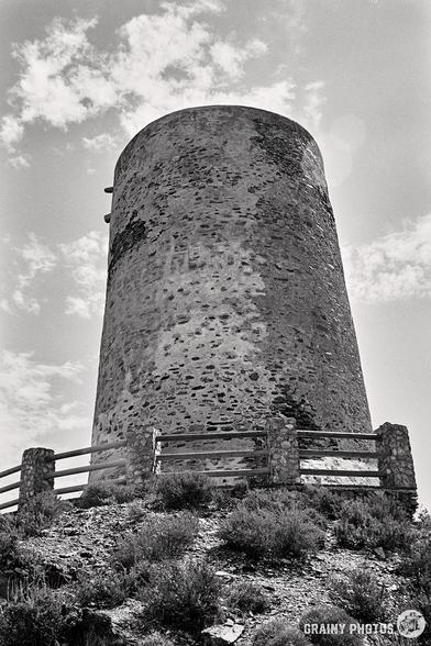 A black and white photograph captures the cylindrical stone tower (Torre Cautor), set against a partly cloudy sky. Thge tower is surrounded by low stone posts and wooden railings and sparse vegetation on a Spanish hillside.