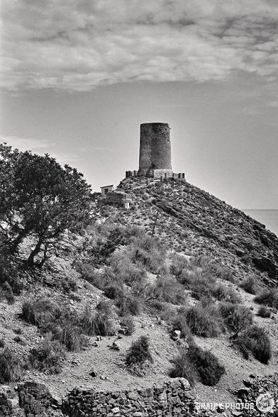 A black-and-white photo captures the stone tower of Torre Cautor atop a rocky hill in Spain, surrounded by sparse vegetation. The cloudy sky enhances its dramatic feel, and the rough terrain in the foreground is dotted with scattered bushes and trees.