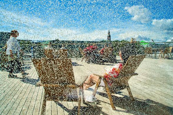 Image taken from behind of two people sitting on chaise long and sunbathing. The day is sunny and bright, blue sky with some clouds at the horizon. Between the subjects and the photographer there's a glass wall covered by thousands and thousands of little golden dots that give the image a dreamy feeling.