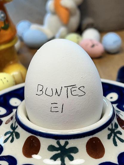 Ein weißes Ei mit der Aufschrift „Buntes Ei“ in einem Eierbecher aus Porzellan. 