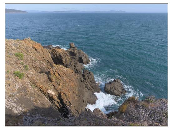 Orange and white lichen adorn rocky cliffs at lower left. Looking steeply down, along a rough coastline to a nearby point, with a plunging drop over the edge in the immediate foreground.  Distant land across a large bay, hazy blue sky in the late afternoon, clear green seas with a small swell.
