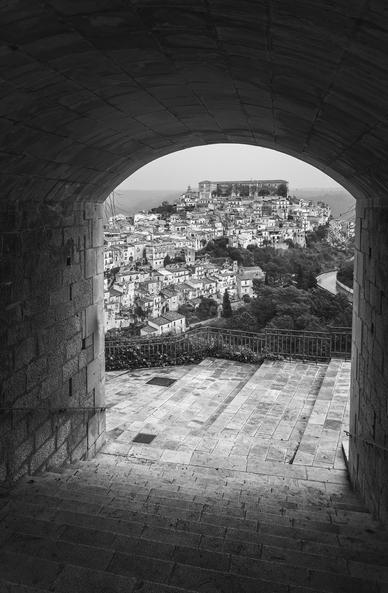 A black and white photo of the old town of Ragusa, Sicily, viewed through a stone archway from a hill across the valley. Steps lead down from the arch down to the valley below. 