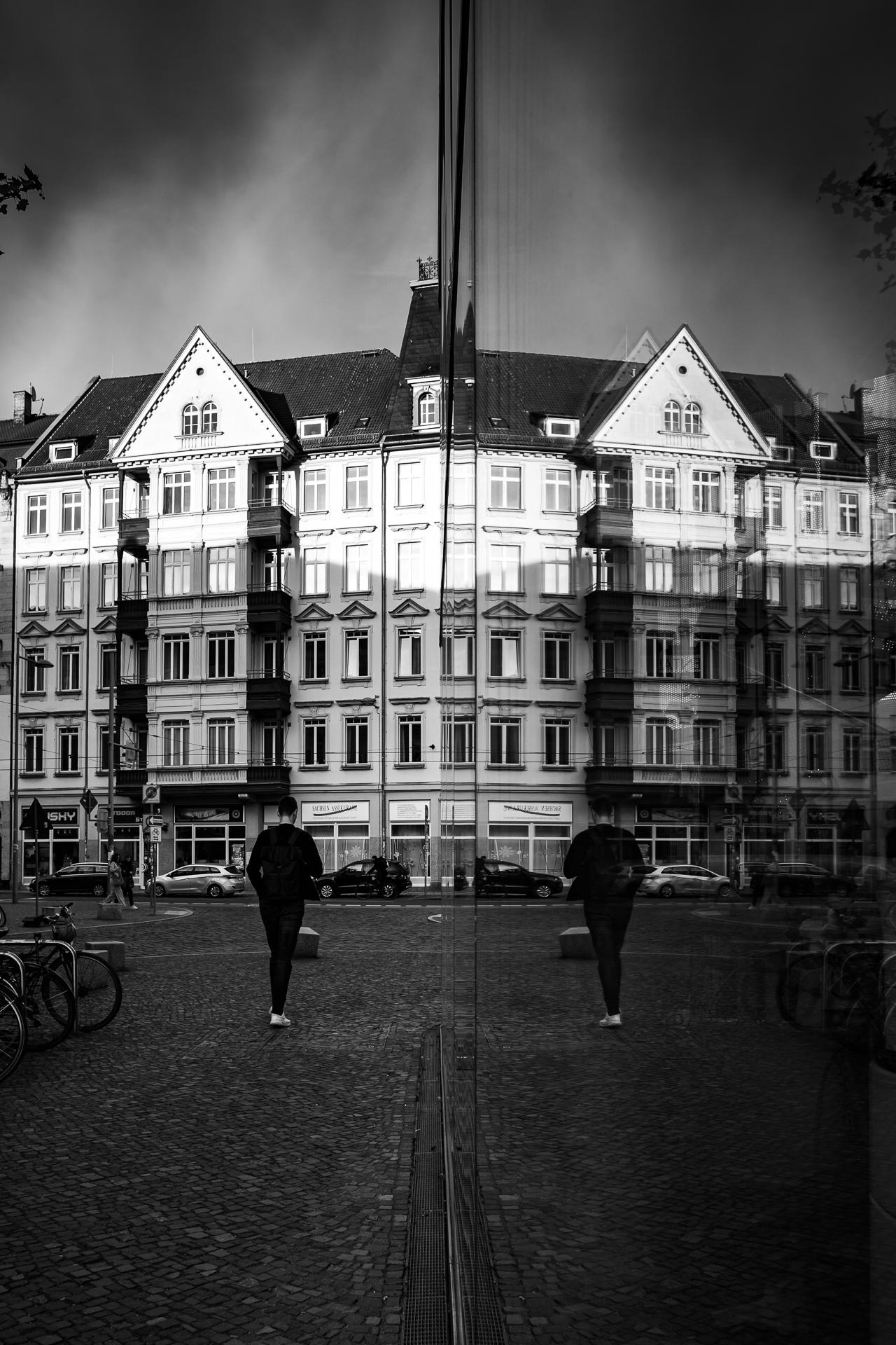 A solitary figure walks across a cobblestone street. The pedestrian is seen from behind, walking away from the viewer. To the side, a large glass window shows a reflection of the person and the buildings he's walking towards. A solitary figure walks across a cobblestone street. The pedestrian is seen from behind, walking away from the viewer. To the side, a large glass window shows a reflection of the person and the buildings he's walking towards.