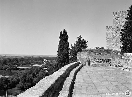 A stone terrace with crenellated walls overlooks a scenic landscape of trees and a distant river. Tall, cyprus trees stand nearby under a clear sky while part of a fortified structure (now a hotel) is visible on the right.