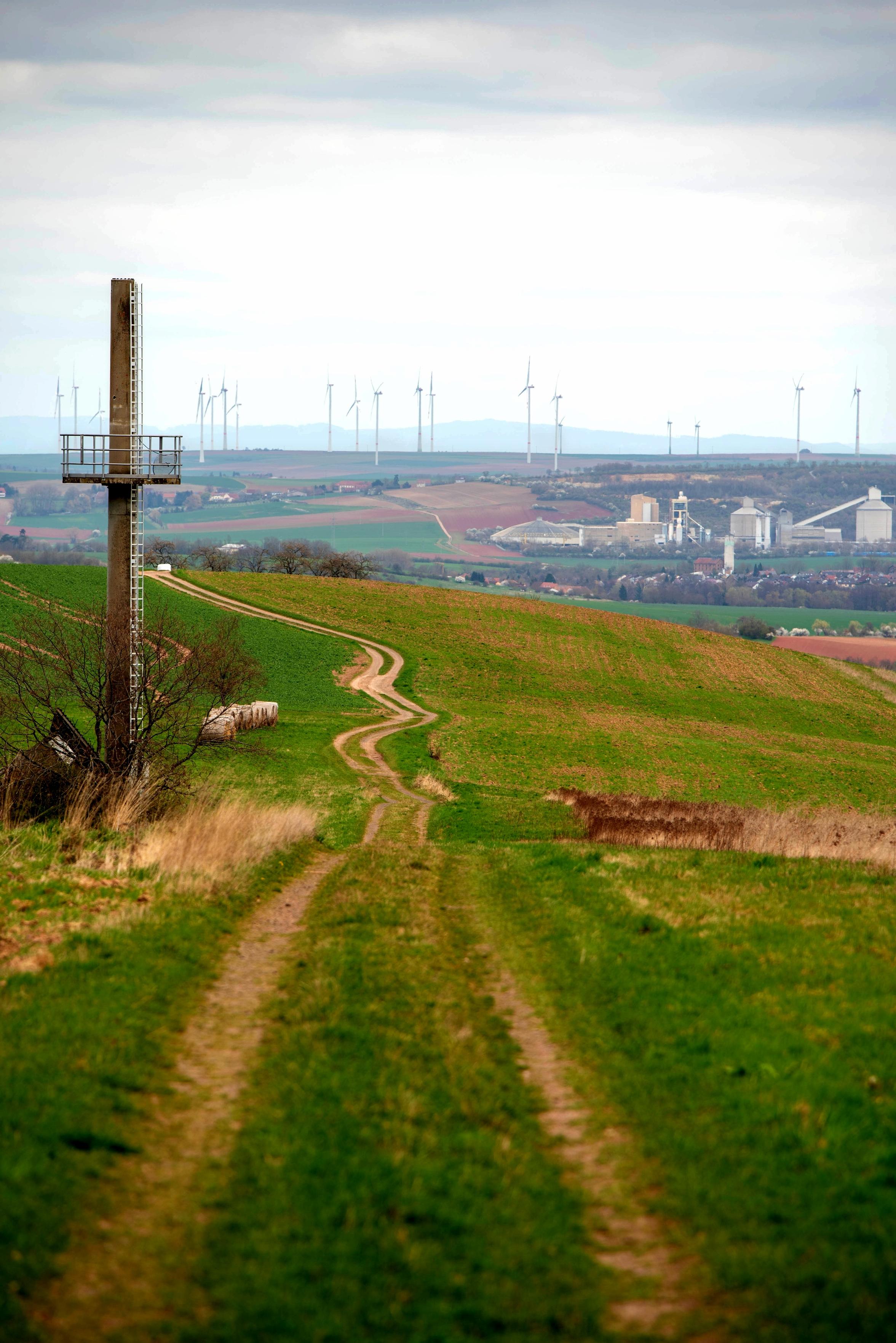 Ein Feldweg der sich durch Felder schlängelt. Links im Bild ein ehemaliger Sendemast. Im Hintergrund am Horizont stehen Windkraftanlagen. Rechts ein Zementwerk. Ein Feldweg der sich durch Felder schlängelt. Links im Bild ein ehemaliger Sendemast. Im Hintergrund am Horizont stehen Windkraftanlagen. Rechts ein Zementwerk.