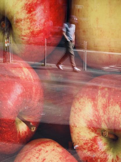 Colour portrait-format image showing the reflection of a man wearing a baseball cap and with his hands in his pockets walking past a window display of giant red apples at a grocery store