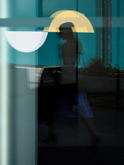 A young woman with long hair is in dark shadows as her reflection is cast onto a plate-glass window which is reflecting the green barriers with yellow and white emblems surrounding redevelopment work in the iconic George Square, Glasgow 