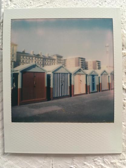 Polaroid photo: A row of small beach huts on a paved surface with multiple coloured doors set against a blue sky.