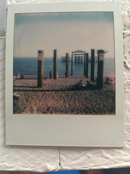 Polaroid photo: The west pier ruins in Brighton. A metal dome like rusted structure out to sea with two rows of rusted pillars on the beach leading towards it. Beach is a pebble beach with people enjoying the hot weather.
