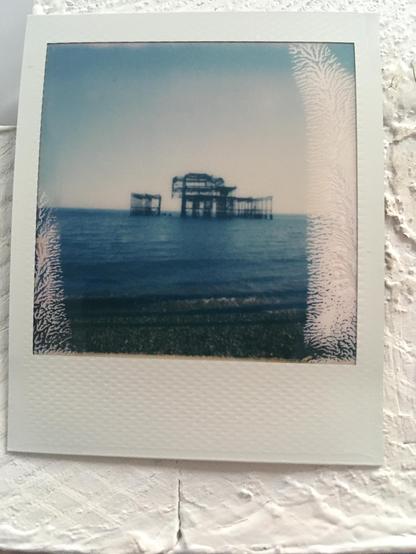 Polaroid photo: The rusted metal remains of the west pier in the glistening blue water of the sea on a clear summers day. The ruins have a noticeable piece missing on the left side where part of the structure stands alone.